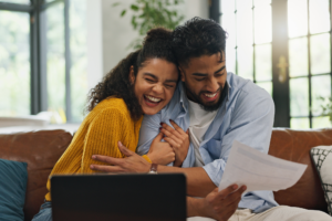 Happy, hug and couple with paperwork, financial planning or excited for home loan on website or app. Laughing, man and woman with document for mortgage, laptop and celebration with embrace in house.